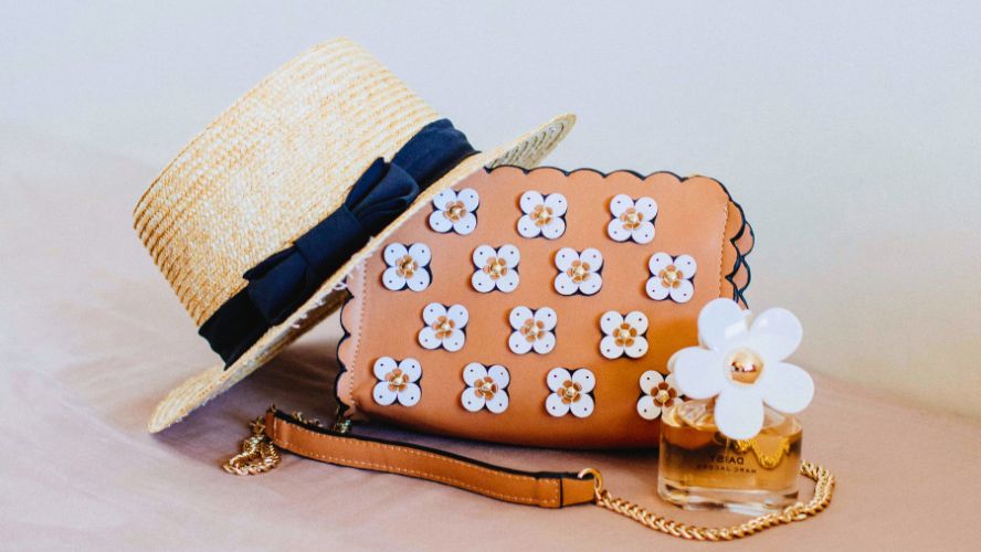 米色花朵裝飾皮包、帶有藏青蝴蝶結的草編平頂帽、Marc Jacobs 小雛菊香水瓶的靜物組合。