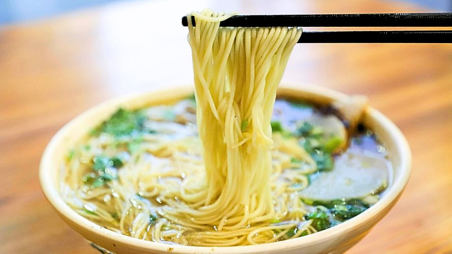 一碗清湯麵，麵條被筷子挑起，湯裡有青菜和其他配菜，呈現中式早餐的經典麵食場景。